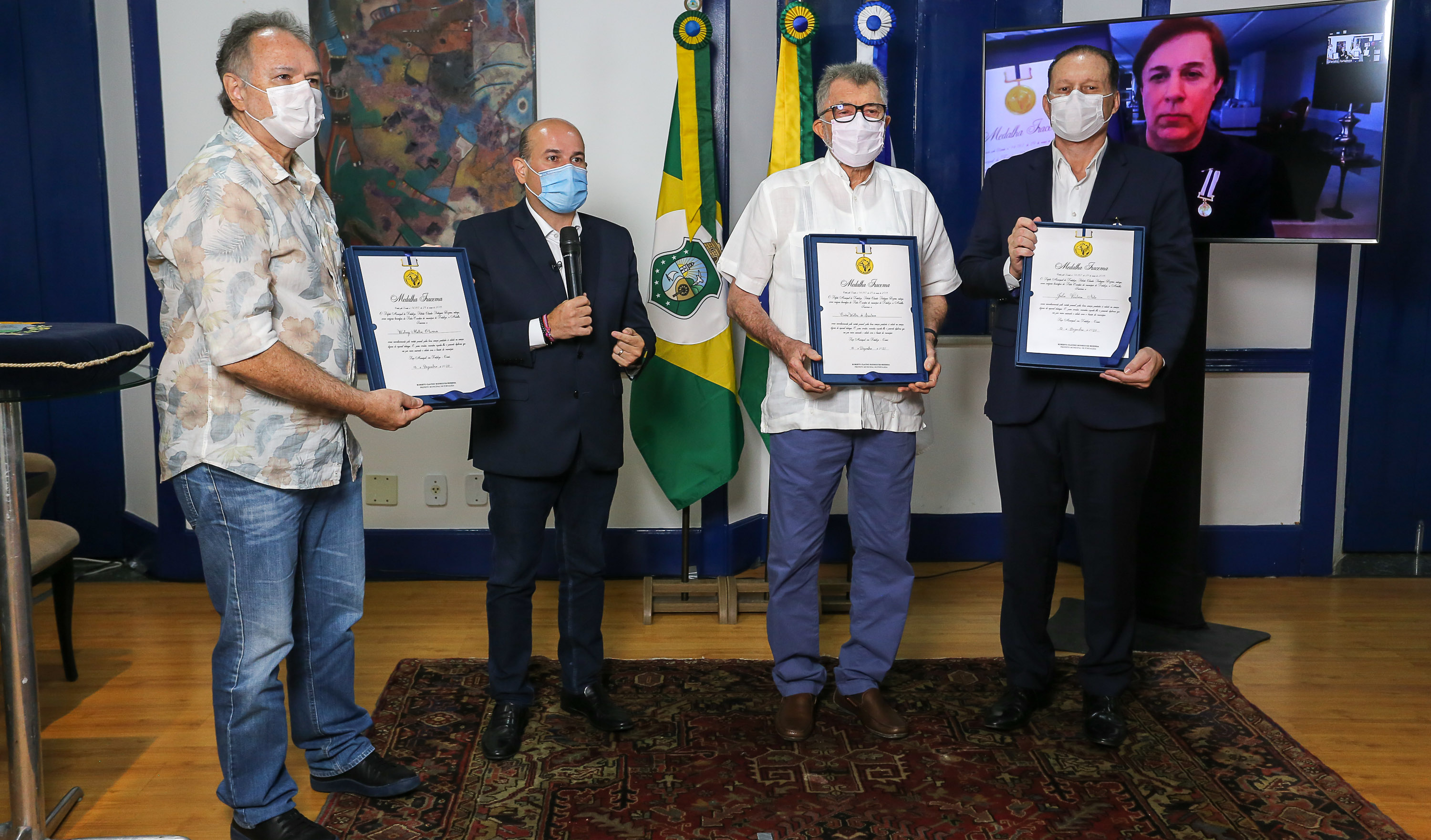 prefeito em pé de máscara ao lado de três homenageados segurando suas medalhas e Tom Cavalcante em TV ao fundo